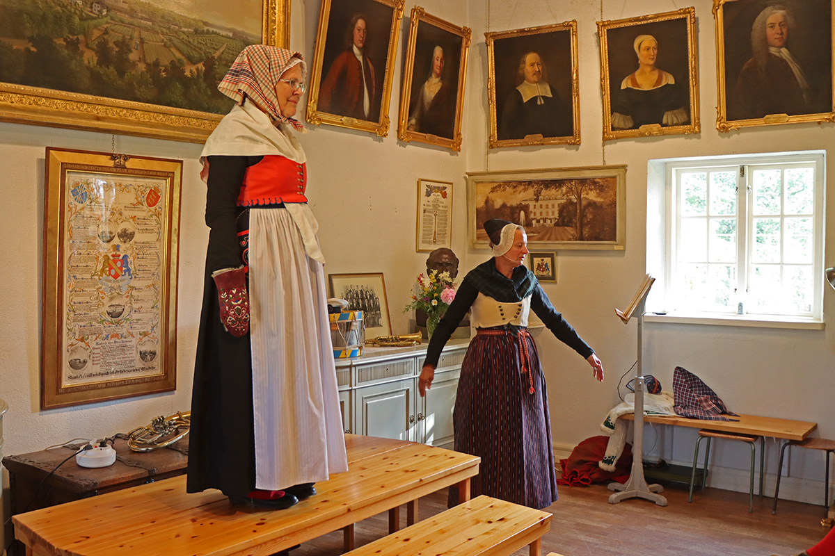 Dräkt för vinter, högsta helg med svart kyrkkjol och jacka, röd fläbblivstycke, vitt förkläde och halskläde. Röda strumpor och skinnhandskar med broderier. Rutigt halskläde ovanpå skarpmössan. Modell Elisabeth Lindholm.