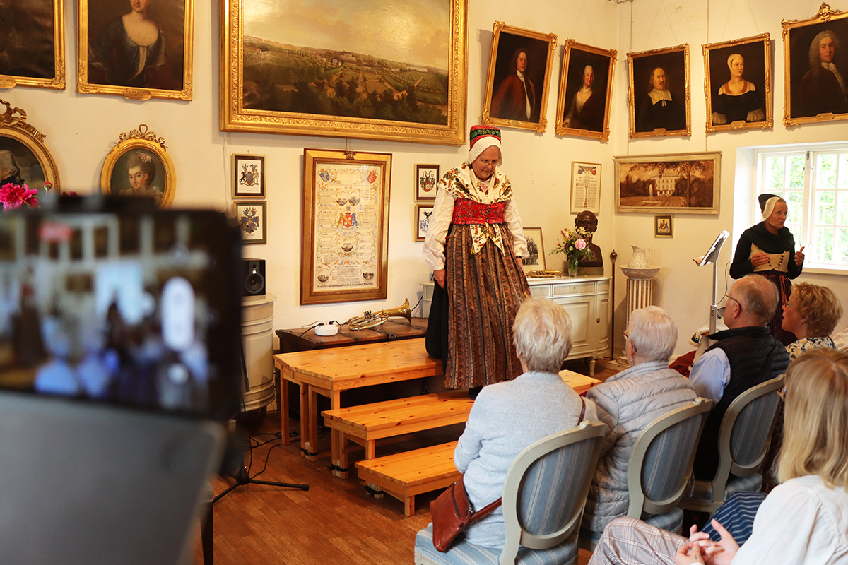 Dräkt för annandag. Svart kyrkkjol, mönstrad bomull, sk kattun i fläbblivstycke, halskläde, förkläde och skarpmössa. Röda strumpor. Modell Elisabeth Lindholm.