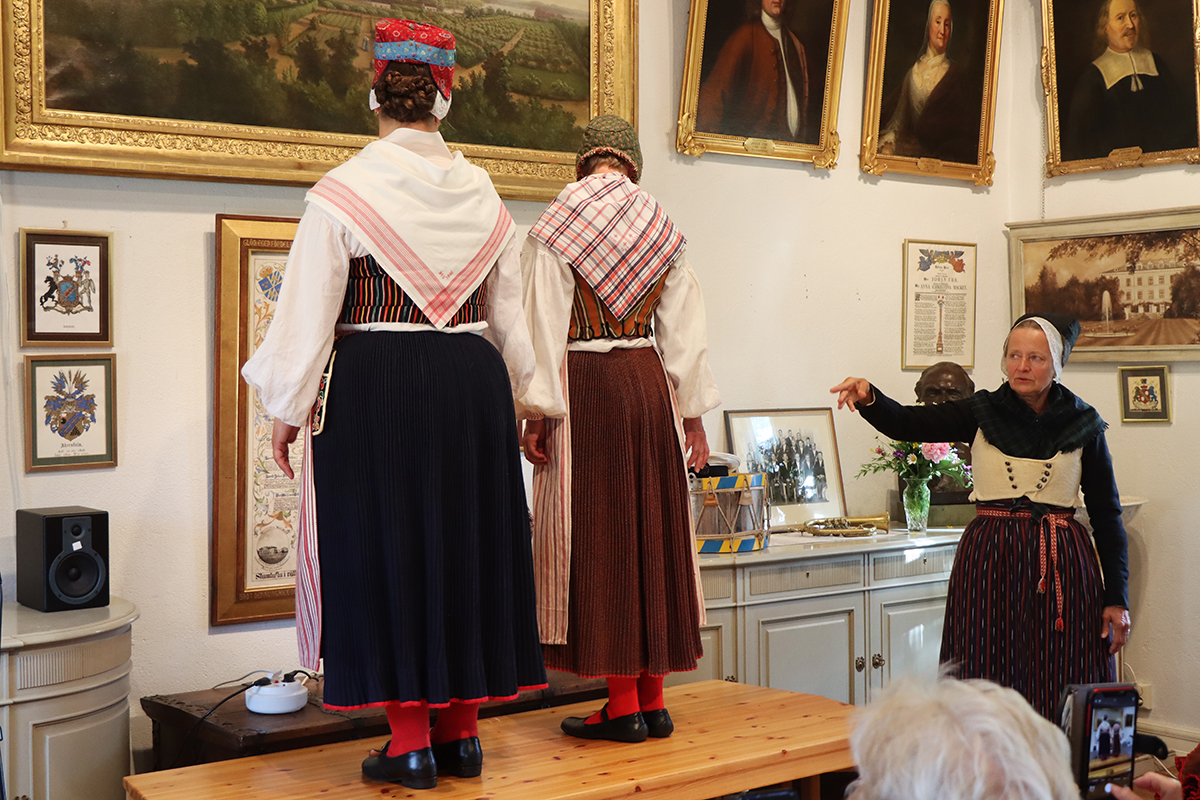 Gamla modets dräkt från slutet av 1700-talet visad bakifrån. Tvinnskört i röd och blå variant, randigt livstycke med skunkliv. Hemvävda förkläden och rutiga halskläden, röda strumpor. Skarpmössa och hylka i kattun. Modeller Susanne Adman och Åsa Palm.