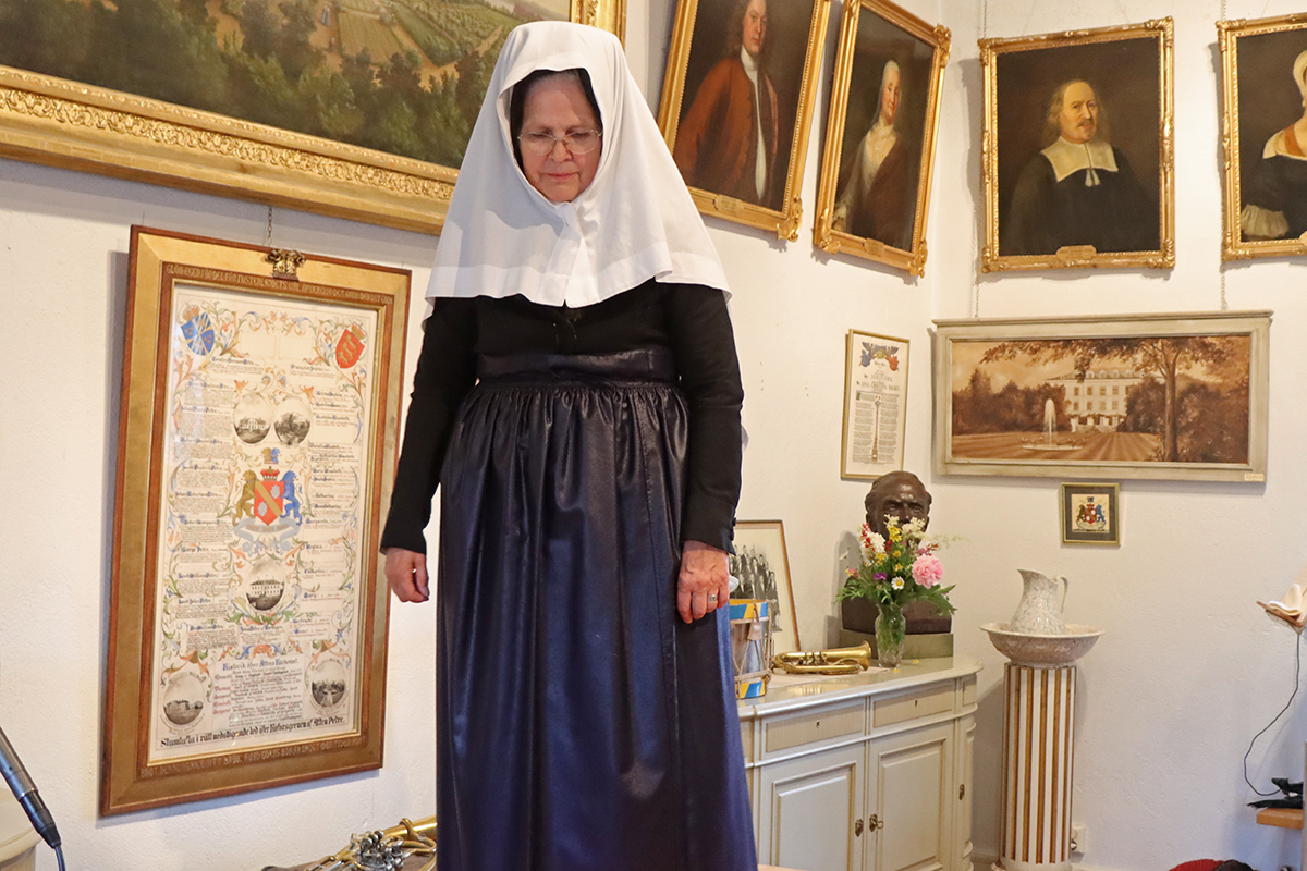 23_2191 Sorgklädsel med svart kyrkjol och jacka. Förkläde i svart rask. Stort huvudkläde i vitt med svart sidenmössa under. Röda strumpor. Modell Elisabeth Lindholm.