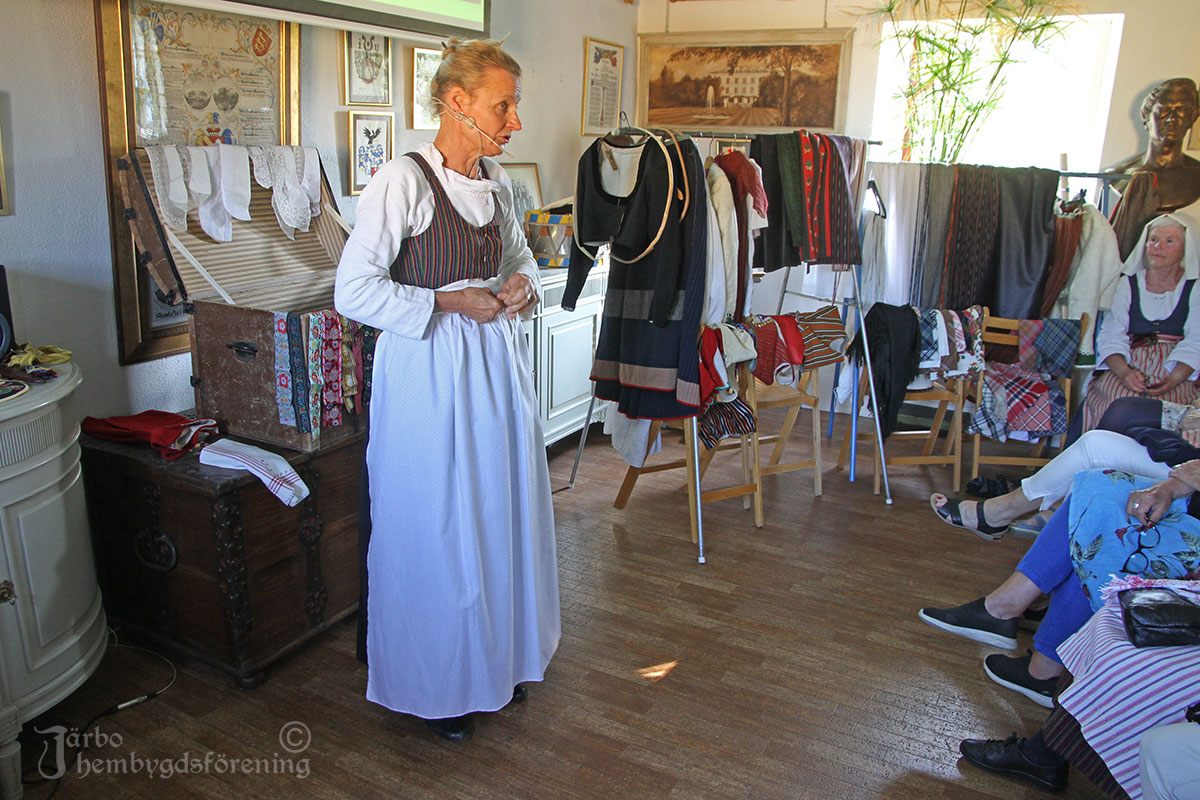 Ann-Mari klär sig inför söndagens besök i kyrkan. Nu kommer det finaste vita köpta kyrkförklädet på. Det är i tunn mönstrad bomull. Under kyrkdräkten är hon redan färdigklädd för söndagseftermiddag i den randiga livkjolen.