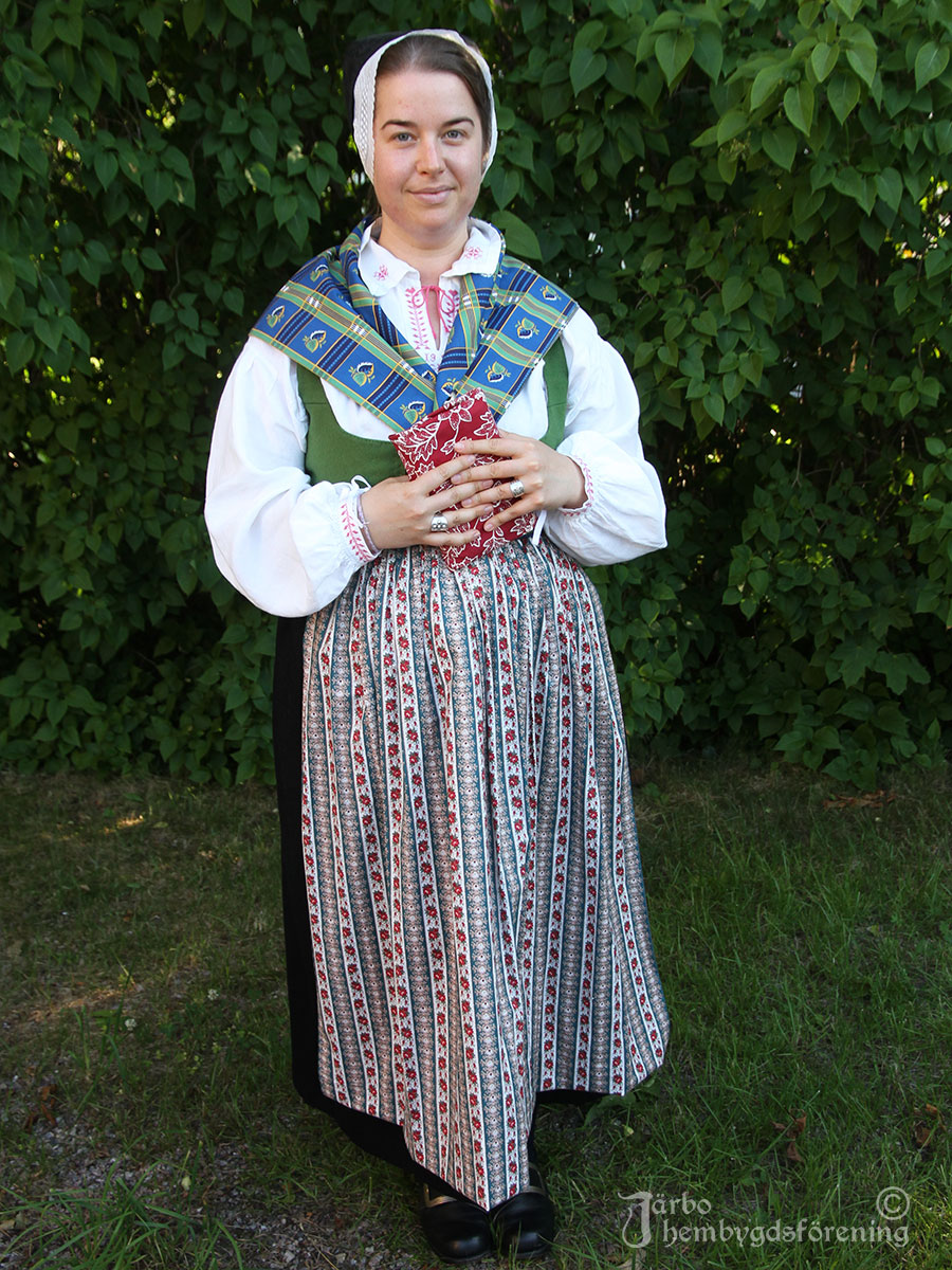 IMG_1855 Dräkt för konfirmation och allvarshelg, Ovansjö socken. Dräkten består av svart kjol, grönt fläbblivstycke, grönblommigt förkläde och halskläde. Finaste svarta bindmössan i siden med vitt stycke. Blåa strumpor. Psalmboken inlindad i ett bokkläde. Modell Sofie Molander.