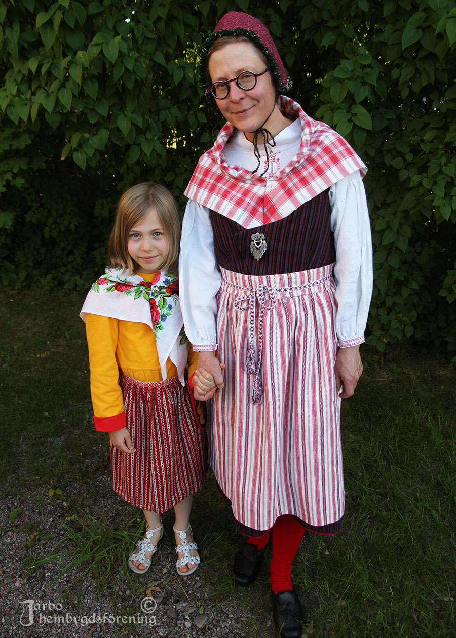 IMG_1899 Åsa Palm i randig livkjol från Ovansjö. Rödrutigt halskläde och rödrandigt kvistförkläde och röda strumpor. På huvudet har hon en hylka för vardagsbruk, mjuk och bekväm. Dotter Svea har en gul Leksandskolt med rödmönstrat förkläde och rosigt halskläde.