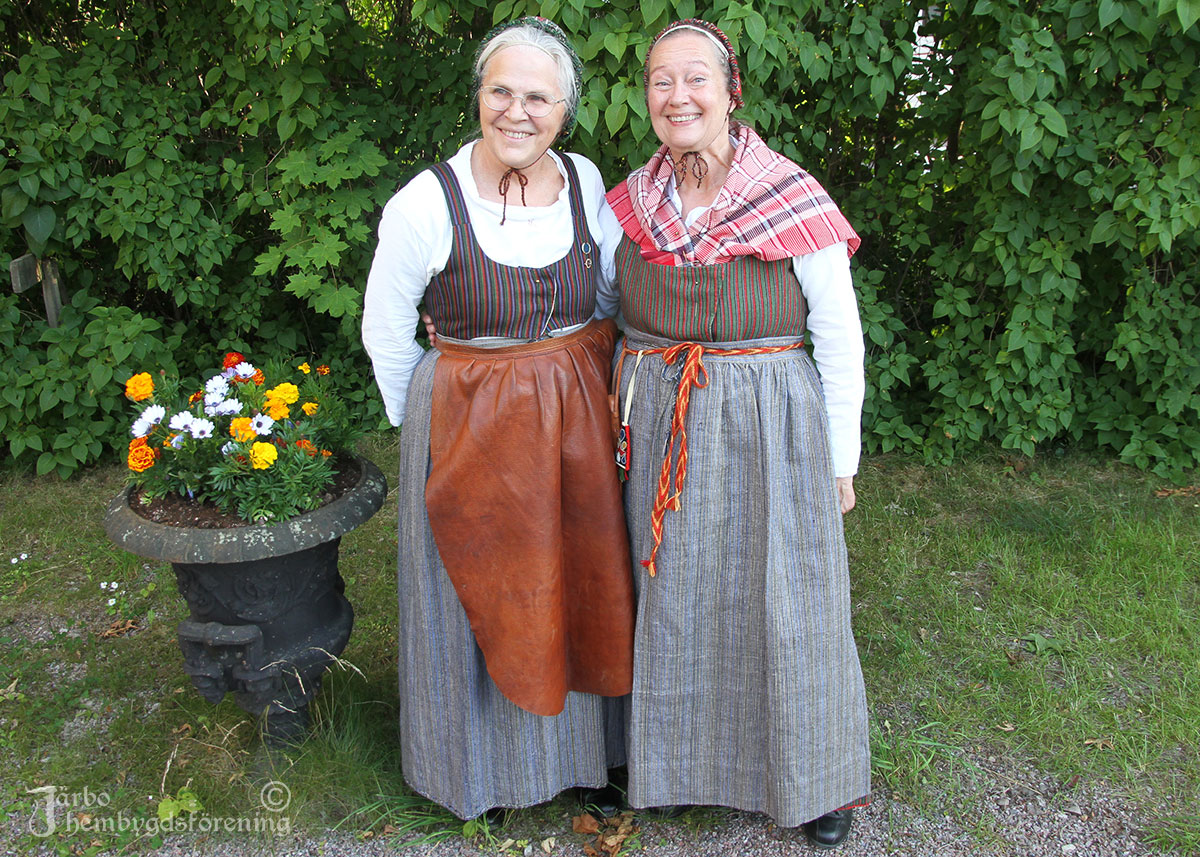 IMG_1943 Elisabeth och Gunilla i vardagsdräkter från Ovansjö. Olika randning på livkjolarna och hemvävda förkläden i lin därtill. Hylkor som huvudbonader. Gunilla har på sig ett flätat livband med ett nålhus. Det dög inte att gå omkring overksam förr i tiden. Elisabeth har även ett förkläde i skinn för arbete. Ofta hade kvinnorna flera kjolar och förkläden på sig, det ena över det andra. Bra mot kylan, det värmde gott.