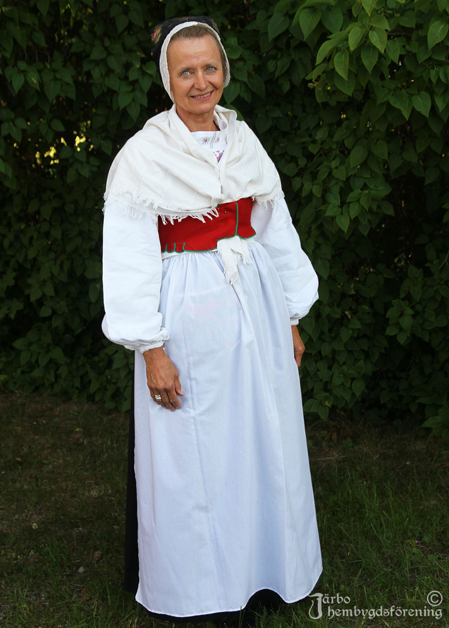 IMG_1945 Ann-Mari Jerner färdigklädd för kyrkbesök för högsta helg, Ovansjö socken. Finaste svarta kjolen, vit broderad överdel, vitt förkläde i köpetyg, rött fläbblivstycke och vitt halskläde med tambursöm. Kjolsäcken sitter under förklädet. Röda sydda strumpor och svarta skor. Finaste skarpmössan med blommigt mössband och broderat stycke.