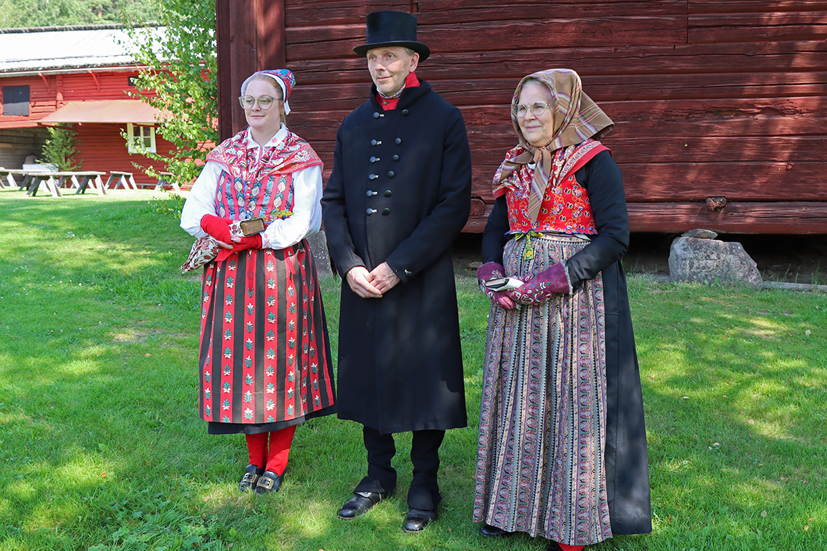 granslos_4160 Från vänster: Tova har en dräkt för annandagar på sommaren i Ovansjö med svart kyrkkjol, vit överdel, blommig kattun i glada färger på livstycket, i förkläde, halskläde och bindmössa. På händerna röda stickade halvvantar. Mikael är klädd för vintern i svart kyrkrock och svart hög hatt, svarta knäbyxor och snösockor, väst i blommig röd kattun. Elisabeth är annandagsklädd för vintern och har lite mörkare kattun i förklädet och huvudkläde, vilket passar en äldre kvinna. Broderade skinnvantar.