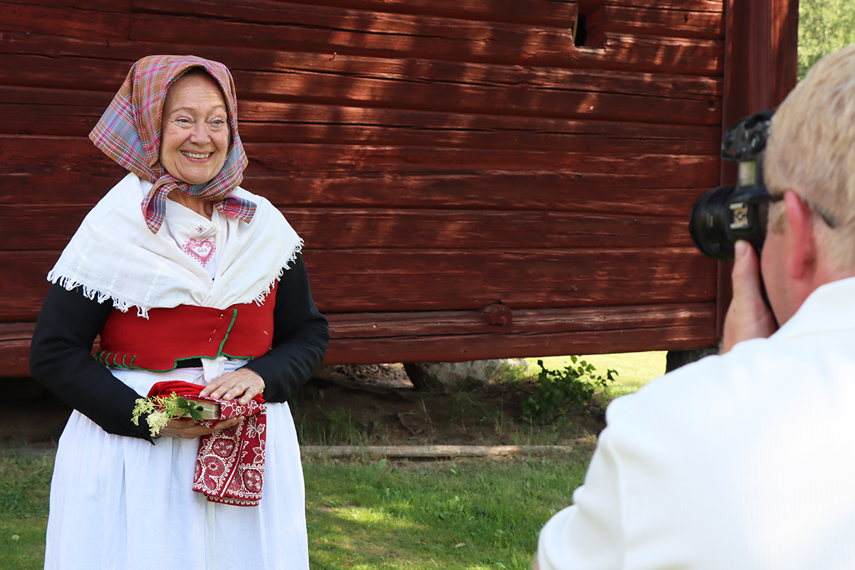 granslos_4169 Gunilla är glad och uppklädd i finaste och högtidligaste kyrkdräkten för vinterbruk. Svart kjol med svart tröja, fin överdel i vitt linne med broderier, tambursömshalskläde och rött fläbblivstycke, som sätts ovanpå den svarta tröjan. Sidenmössa med fint tyllborderat stycke under det rutiga halsklädet. Röda halvvantar och psalmboken inlindad i ett bokkläde av kattun.