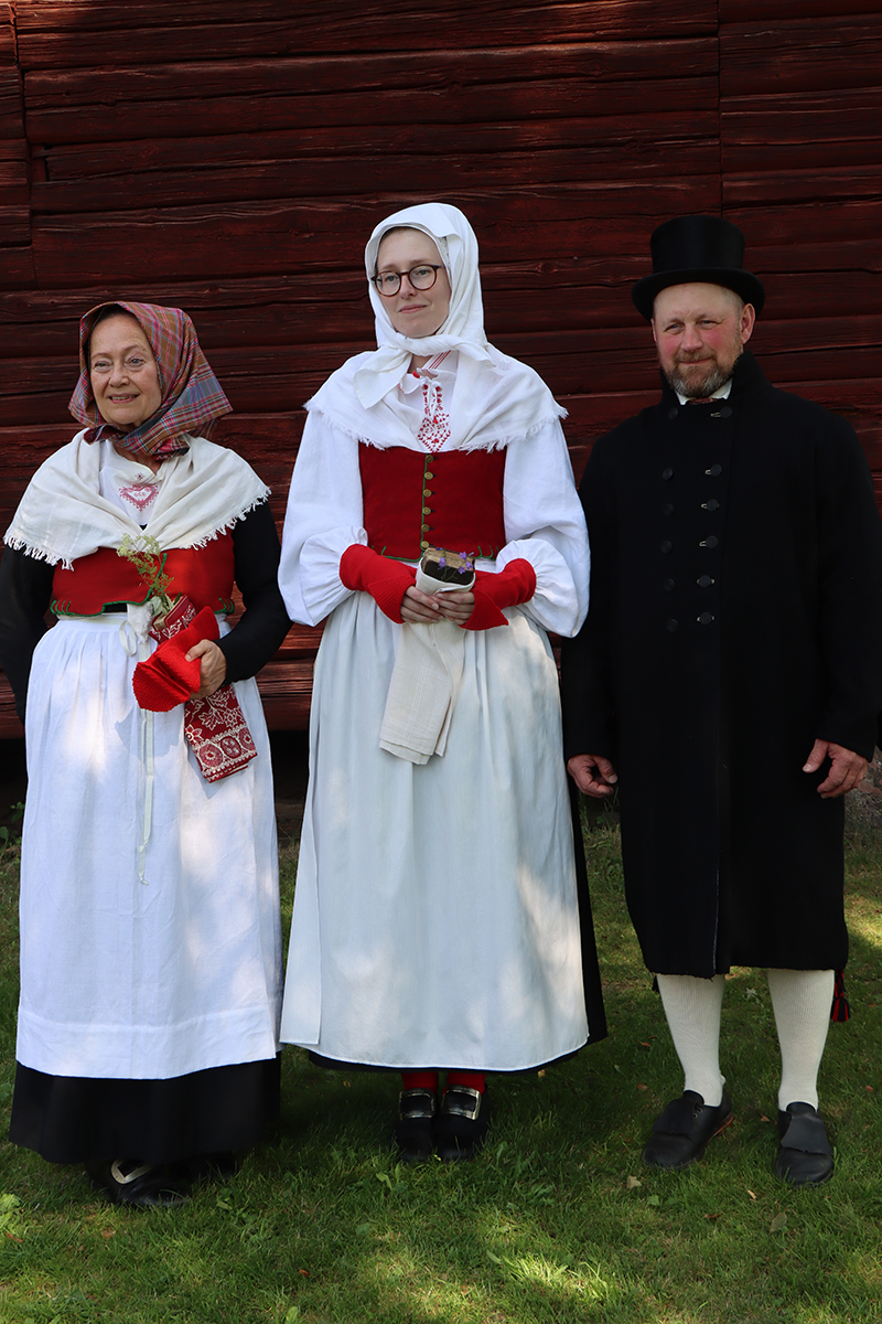 granslos_4191 Gunilla i högsta helgdräkt för vinter står bredvid Madelene som är klädd på samma sätt fast för sommar. Det som skiljer kläderna åt är den svarta tröjan som Gunilla satt under livstycket. Robert är stilig i svart kyrkrock, hög hatt, randig väst och vita strumpor.
