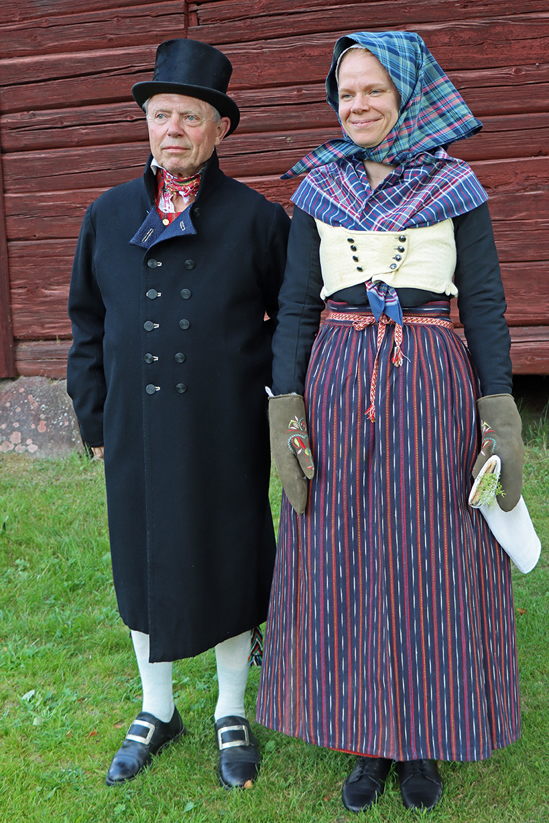 granslos_4204 Bengt är klädd för högsta helg i svart kyrkrock, hög hatt, rödrandig hemvävd väst och rött kattunhalskläde. Vita strumpor till. Moa är klädd för vintern och allvarshelg med svart kyrkkjol, hemvävt randigt förkläde, skinnlivstycke med svart tröja under. Skinnvantar. Rutigt halskläde och huvudkläde.