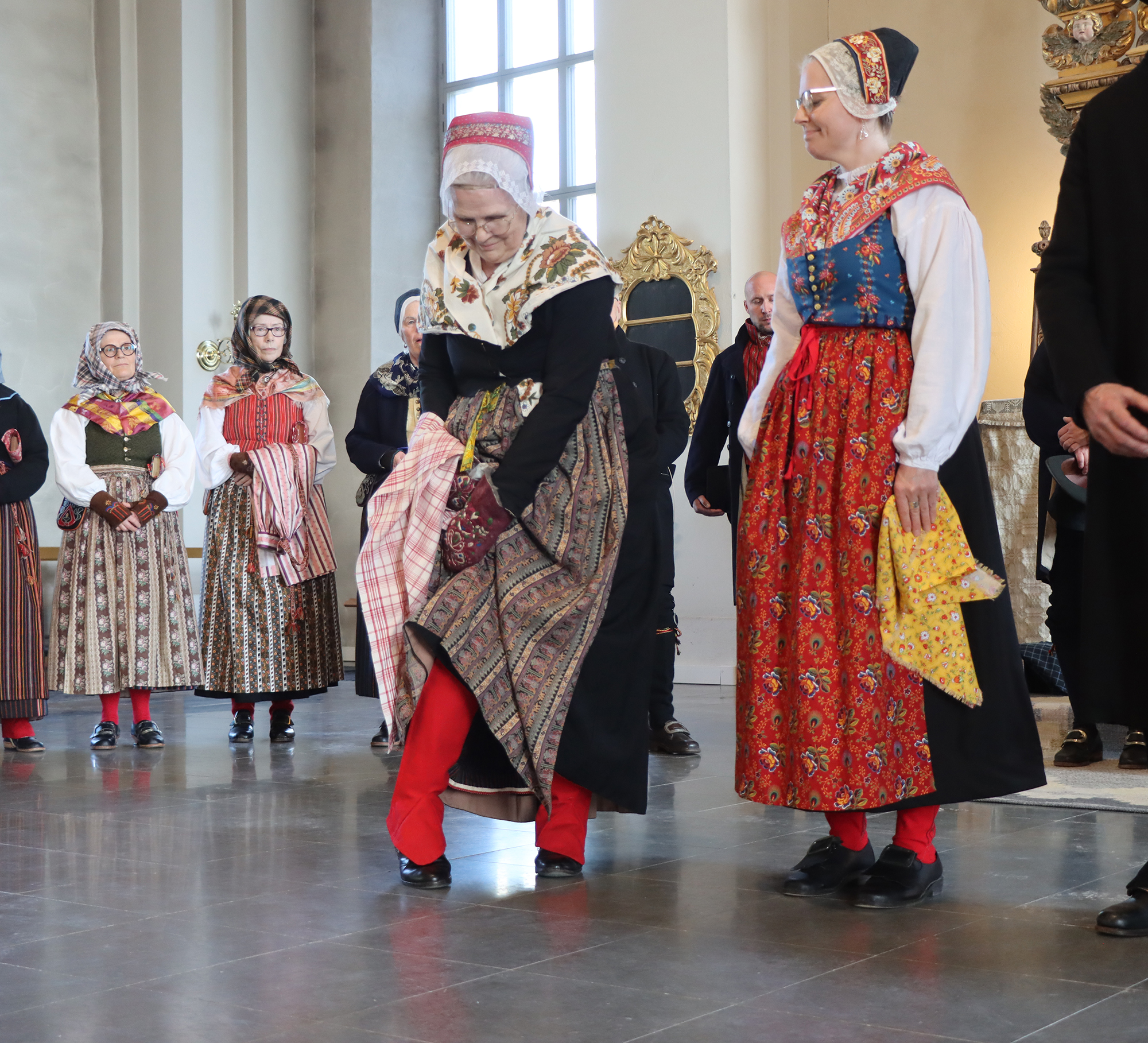 IMG_4816 Elisabeth, klädd för annandagar vintertid, visar upp sina röda snösockor. Susanne bär dräkt för annandagar sommartid. Båda har svarta kyrkkjolar och livstycken, halskläden samt förkläden i kattun. Elisabeth har dessutom en svart kyrktröja över särken. Till den här varianten av dräkt används bindmössor i kattun i olika färger.