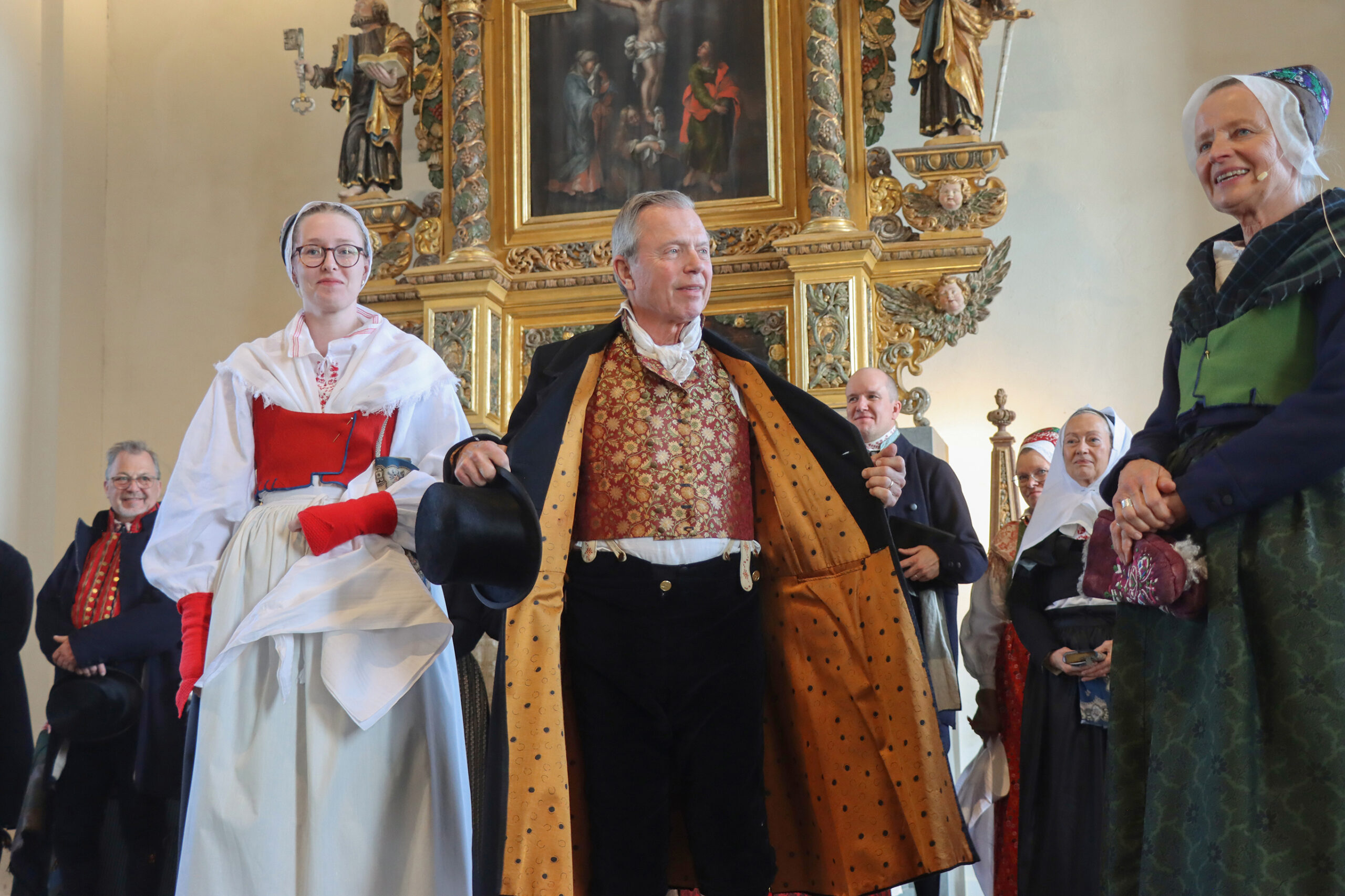 IMG_4840 Madelene är klädd för högsta helg med rött livstycke i yllebrokad, svart kyrkkjol, förkläde och halskläde av fin bomull, stickade snibbhandskar och svart skarpmössa med tumbursöm. Huvudklädet skulle vara vitt. Bengt är klädd för högsta helg med sidenväst, svarta byxor och långrock.